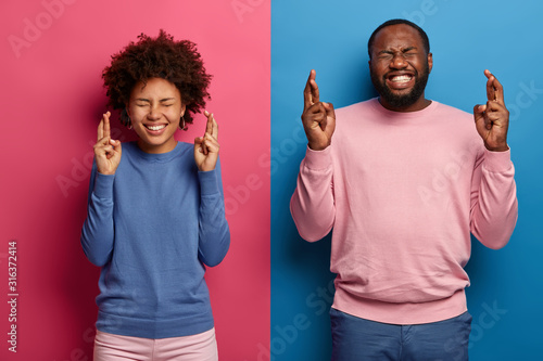 Happy black students cross fingers wait for examination results, pray to get best grade, smile broadly, wear casual jumpers isolated over blue and pink background smile toothily. Body language concept photo