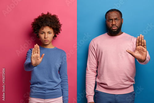 Prohibition symbol. Serious displeased black man and woman make stop gesture with palms, look in dissatisfaction, wear casual clothes, disagree with something, isolated over blue and rosy studio wall