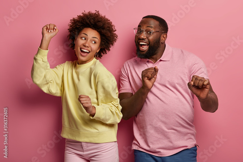 Portrait of energized dark skinned couple dance happily, have inspired joyful face expressions, smile broadly, moves with music, clench fists and raise arms, isolated over rosy pastel background