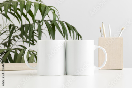 Two mugs mockup with workspace accessories on a white table and a palm plant.