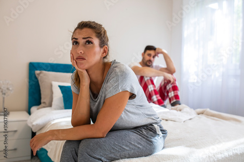 Young upset girl sitting on the edge of the bed, against her boyfriend, lying in bed. Frustrated sad girlfriend sit on bed think of relationship problems