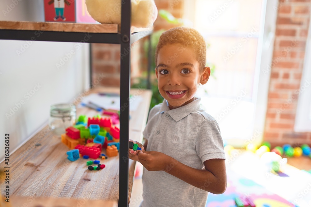 Beautiful african american toddler standing playing with small building blocks on shelving at kindergarten - obrazy, fototapety, plakaty 
