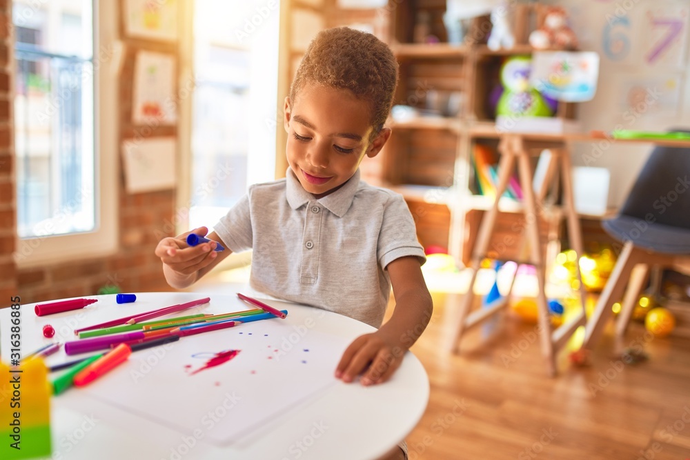 Beautiful african american toddler drawing using paper and marker pen smiling at kindergarten