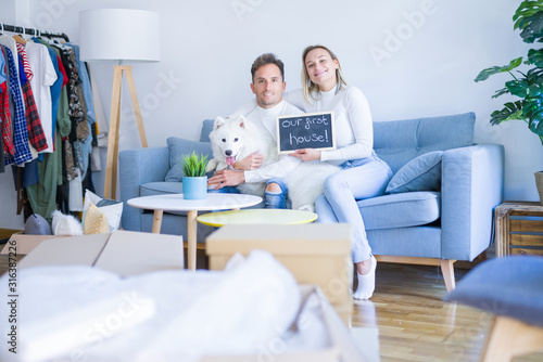 Young beautiful couple with dog sitting on the sofa holding blackboard with message at new home around cardboard boxes © Krakenimages.com