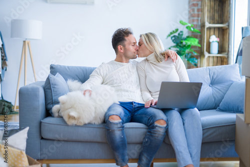 Young beautiful couple with dog sitting on the sofa kissing using laptop at new home around cardboard boxes