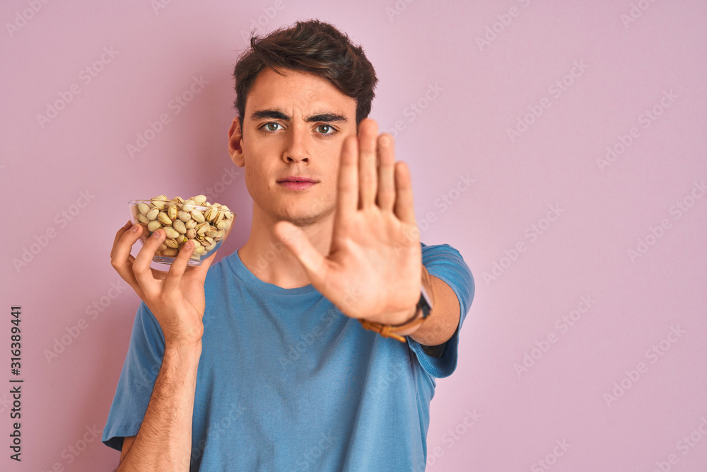 Teenager boy holding a bunch of pistachios over isolated pink background with open hand doing stop sign with serious and confident expression, defense gesture Teenager boy holding a bunch of pistachios over isolated pink background with open hand doing stop sign with serious and confident expression, defense gesture