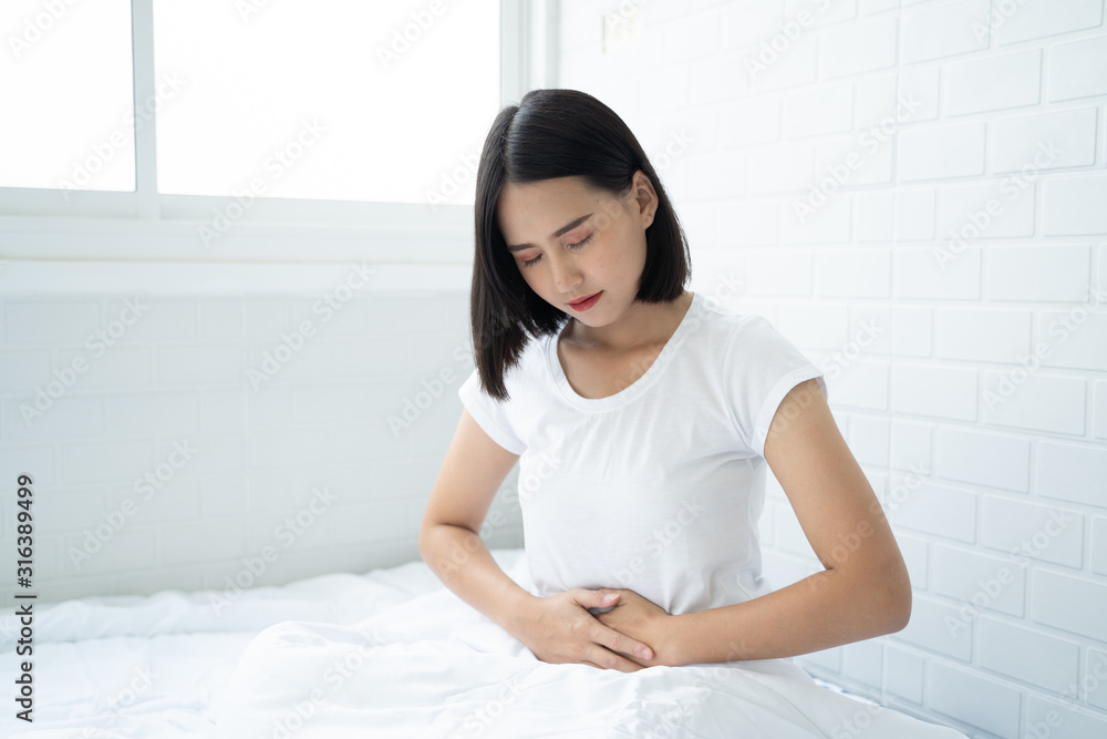 Asian woman unhappy lying on the bed looking sick, suffers from stomach ache in the bedroom, stomachache because of menstruation and eating spoiled food, Chronic gastritis. Abdomen bloating concept Asian woman unhappy lying on the bed looking sick, suffers from stomach ache in the bedroom, stomachache because of menstruation and eating spoiled food, Chronic gastritis. Abdomen bloating concept