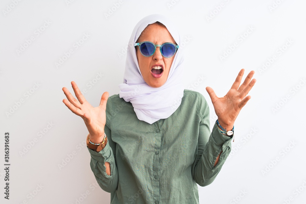 Young Arab woman wearing hijab and summer sunglasses over isolated background crazy and mad shouting and yelling with aggressive expression and arms raised. Frustration concept. Young Arab woman wearing hijab and summer sunglasses over isolated background crazy and mad shouting and yelling with aggressive expression and arms raised. Frustration concept.