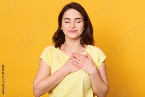 Принти на полотні Image of satisfied generous brunette woman with charming smile, keeps both palms on chest, wearing casual yellow t shirt, standing against studio wall, being kind hearted