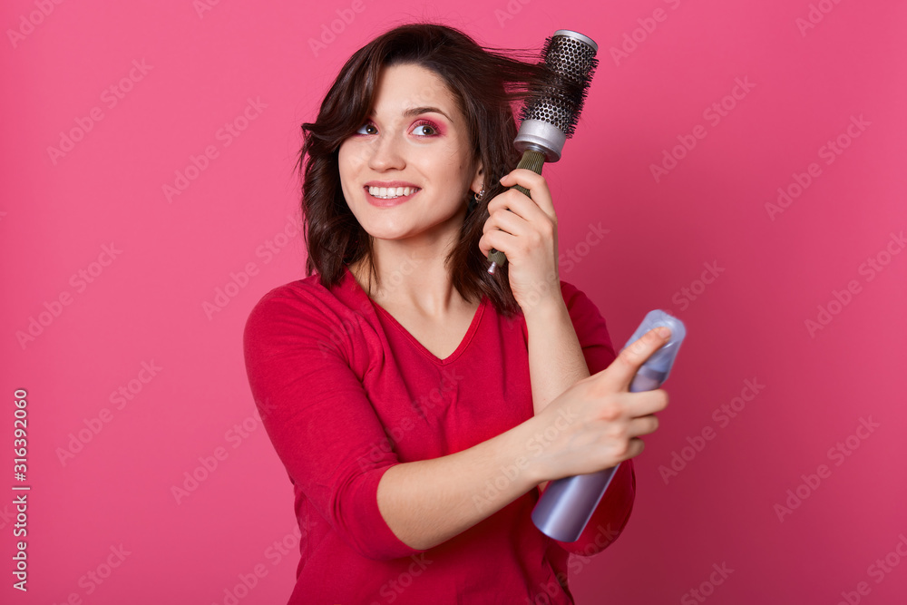 Close up portrait of smiling female hairdresser holding hair brush and hair spray in hands, making hairstyling, brunette being ready for going to work or date, posing isolated over rose background. Close up portrait of smiling female hairdresser holding hair brush and hair spray in hands, making hairstyling, brunette being ready for going to work or date, posing isolated over rose background.