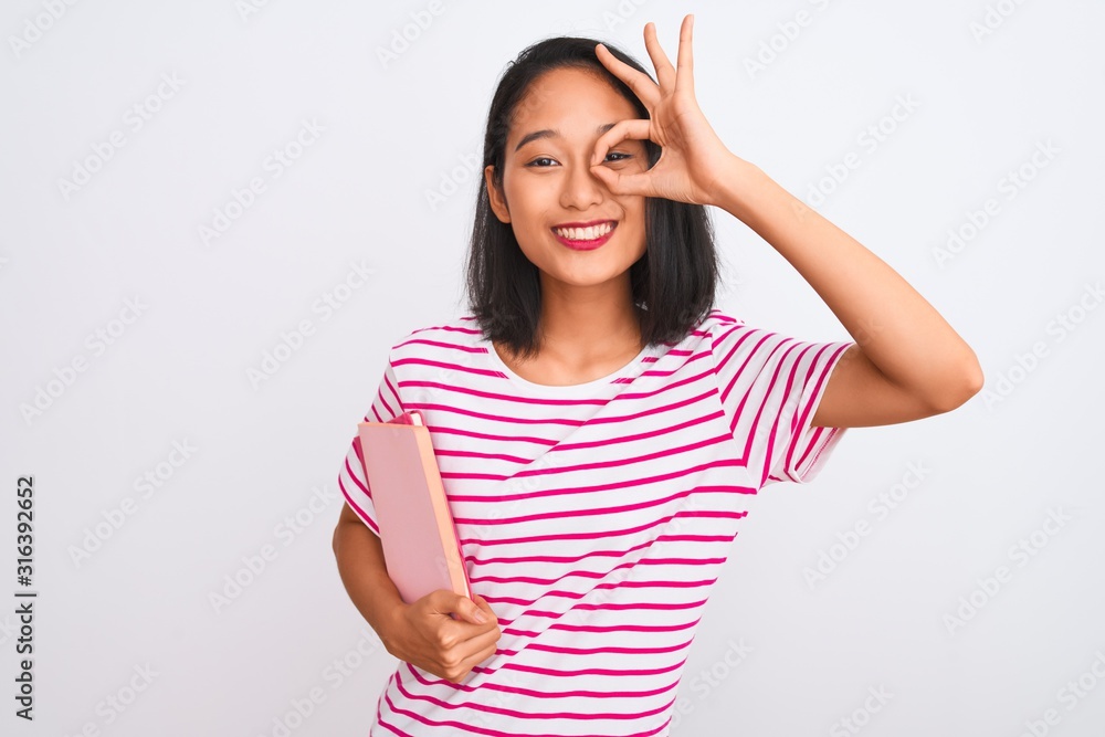Young beautiful chinese woman holding books standing over isolated white background with happy face smiling doing ok sign with hand on eye looking through fingers Young beautiful chinese woman holding books standing over isolated white background with happy face smiling doing ok sign with hand on eye looking through fingers