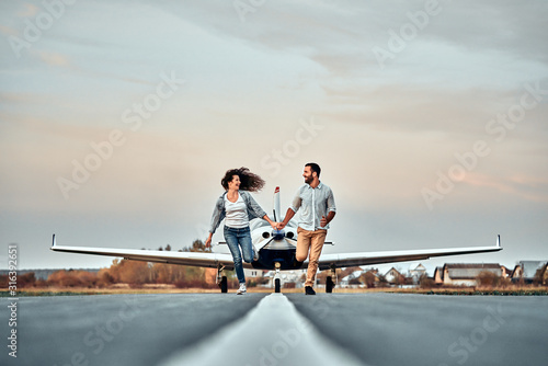 Beautiful romantic couple is holding hands, looking at each other and smiling while running on take-off ground near the aircraft