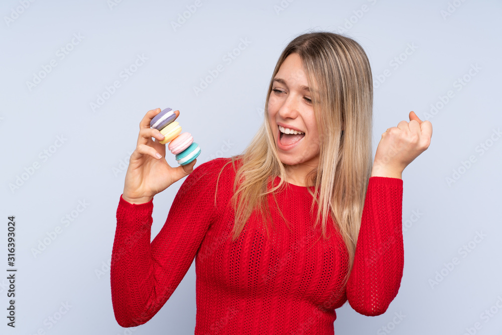 Obraz premium Young blonde woman over isolated blue background holding colorful French macarons and celebrating a victory
