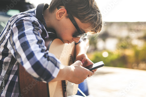 Young boy playing acoustic guitar sitting outdoors. Musician searching music sheet online with smart phone Teen learning to play musical instrument watching Internet tutorial. Educational tech concept