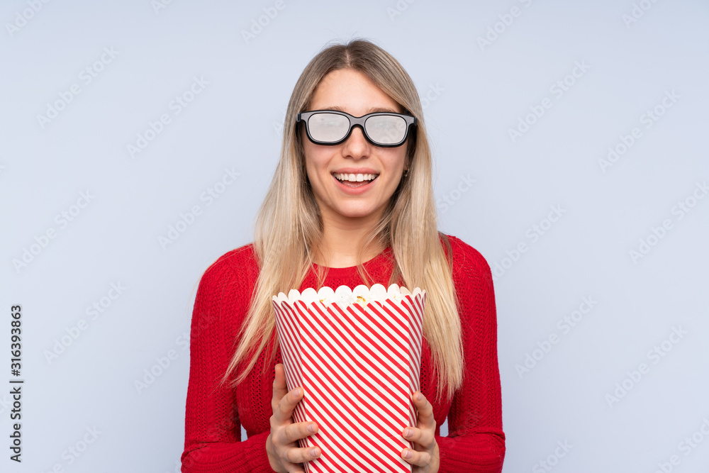 Young blonde woman over isolated blue background with 3d glasses and holding a big bucket of popcorns Young blonde woman over isolated blue background with 3d glasses and holding a big bucket of popcorns