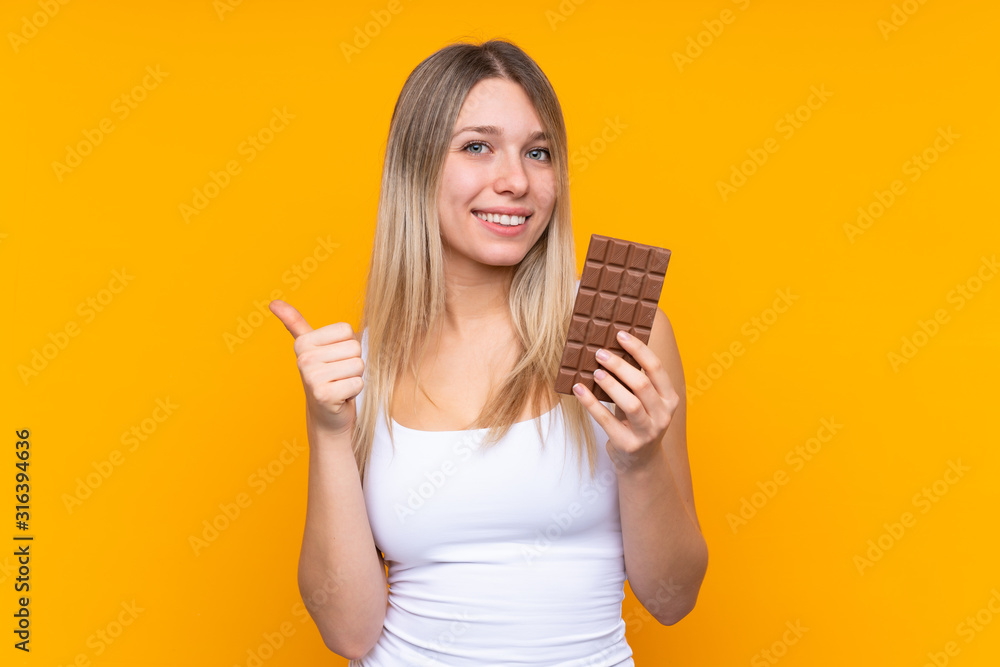 Young blonde woman over isolated blue background taking a chocolate tablet and with thumb up Young blonde woman over isolated blue background taking a chocolate tablet and with thumb up