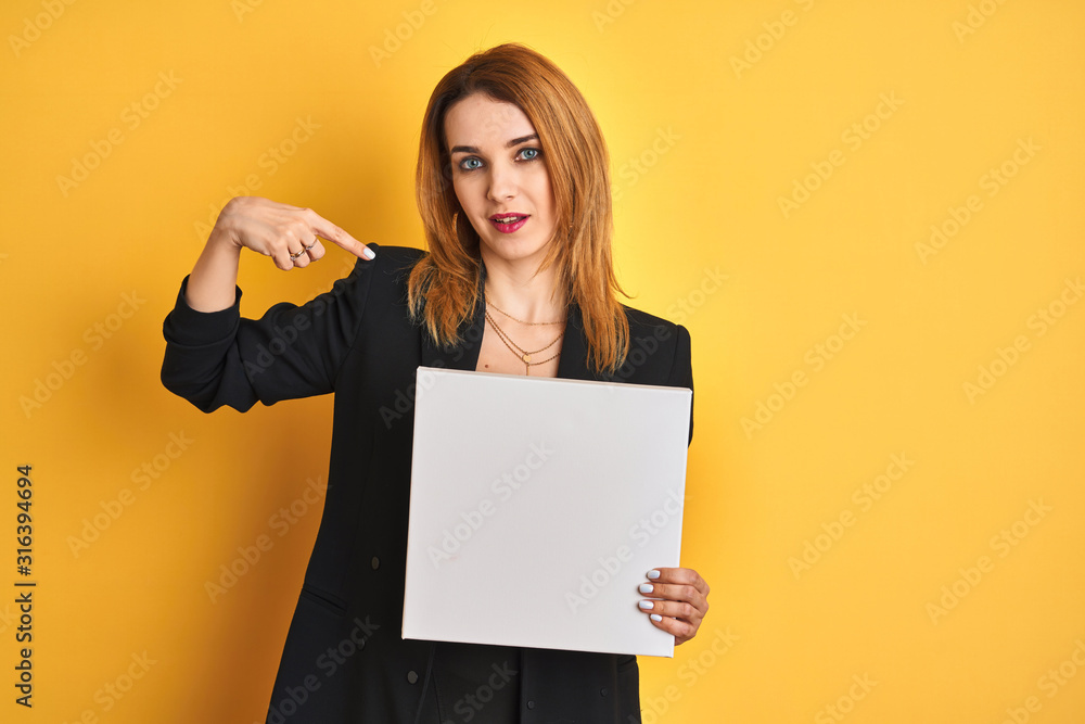 Naklejka premium Young redhead businesswoman with clear eyes holding banner over isolated yellow background with surprise face pointing finger to himself