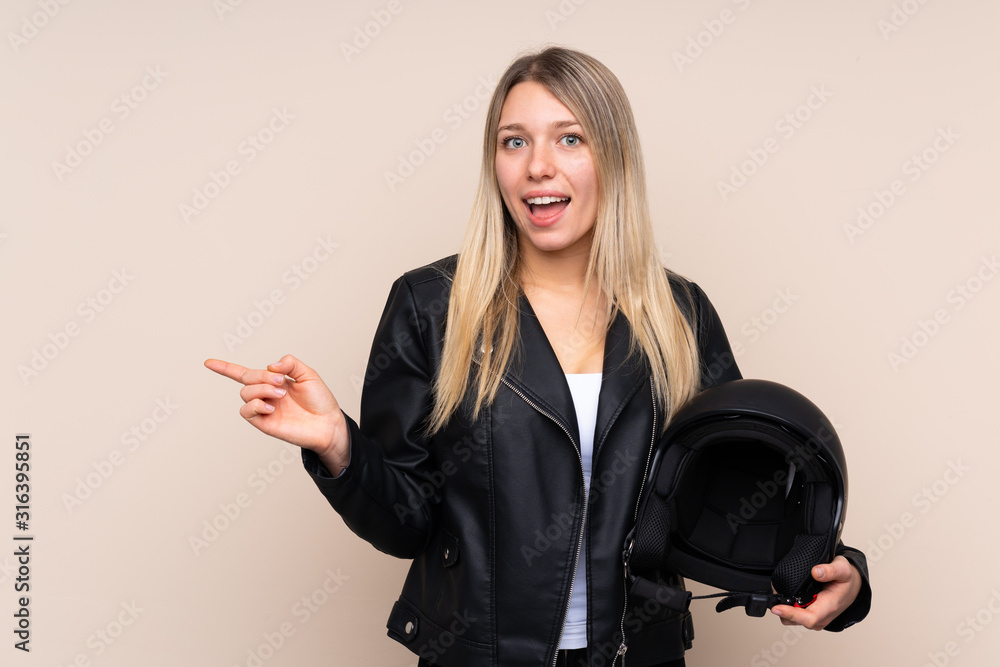 Young blonde woman with a motorcycle helmet over isolated background surprised and pointing finger to the side Young blonde woman with a motorcycle helmet over isolated background surprised and pointing finger to the side