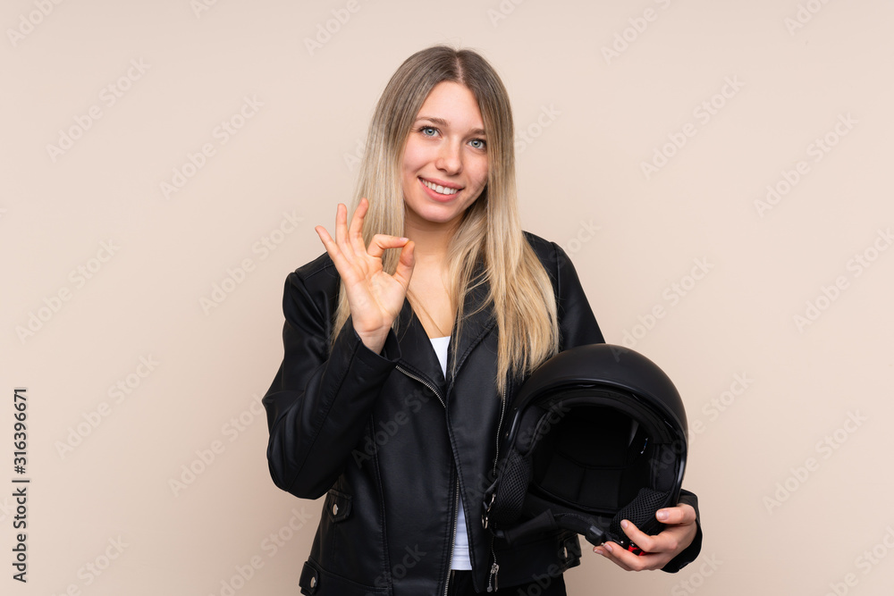 Young blonde woman with a motorcycle helmet over isolated background showing an ok sign with fingers Young blonde woman with a motorcycle helmet over isolated background showing an ok sign with fingers