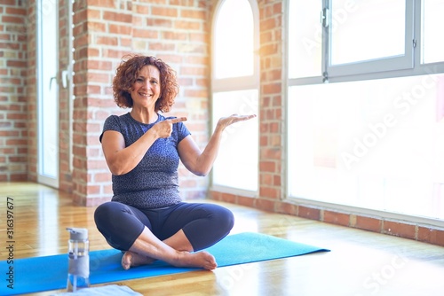 Middle age beautiful sportswoman wearing sportswear sitting on mat practicing yoga at home amazed and smiling to the camera while presenting with hand and pointing with finger.