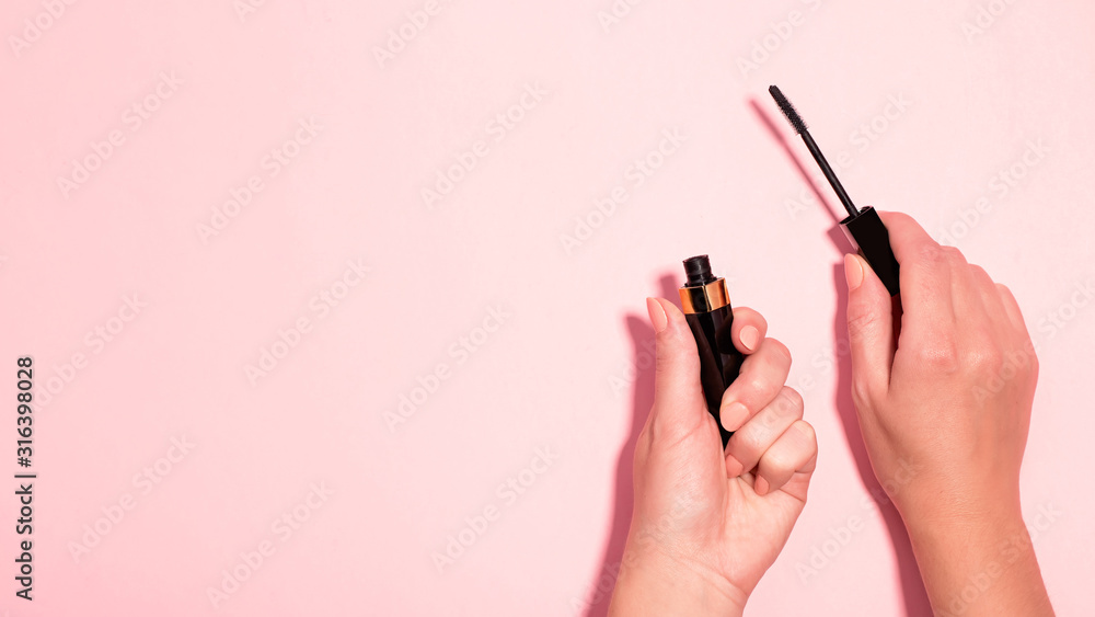 Woman hands holding open black mascara on pastel pink background, top view. Minimal beauty background with makeup products, flat lay, copy space. Eye make up concept Woman hands holding open black mascara on pastel pink background, top view. Minimal beauty background with makeup products, flat lay, copy space. Eye make up concept