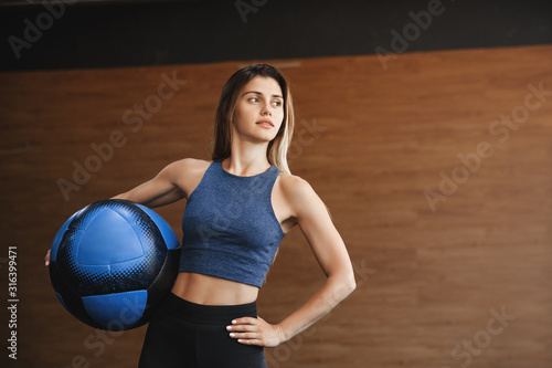 Motivated  assertive good-looking female athlete  fitness instructor in gym wear activewear  hold medicine ball near hip  hold hand waist and turn aside with bossy look  workout functional training
