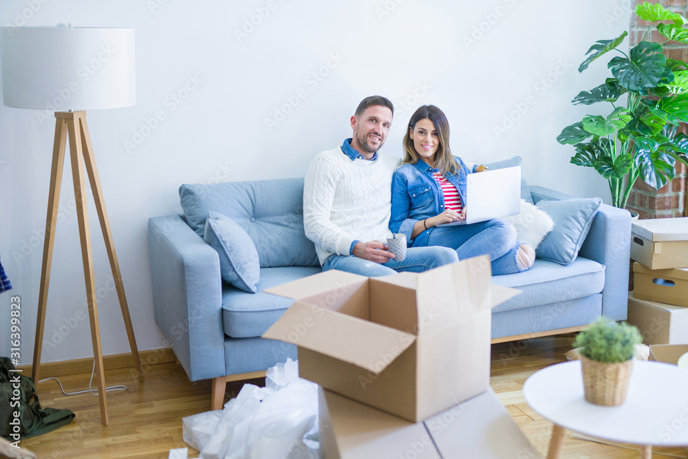Young beautiful couple sitting on the sofa drinking cup of coffee using laptop at new home around cardboard boxes Young beautiful couple sitting on the sofa drinking cup of coffee using laptop at new home around cardboard boxes