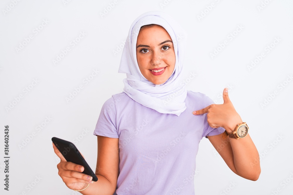 Young beautiful arabian girl wearing hijab using smartphone over isolated white background with surprise face pointing finger to himself Young beautiful arabian girl wearing hijab using smartphone over isolated white background with surprise face pointing finger to himself