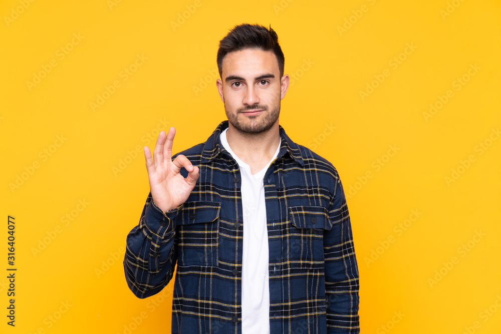 Young handsome man over isolated yellow background showing an ok sign with fingers Young handsome man over isolated yellow background showing an ok sign with fingers