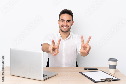 Young business man with a mobile phone in a workplace smiling and showing victory sign