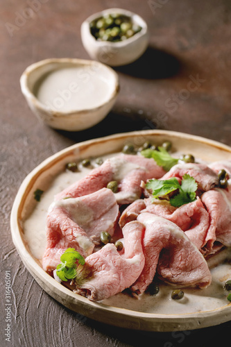 Vitello tonnato italian dish. Thin sliced veal with tuna sauce, capers and coriander served on ceramic plate over dark texture background.