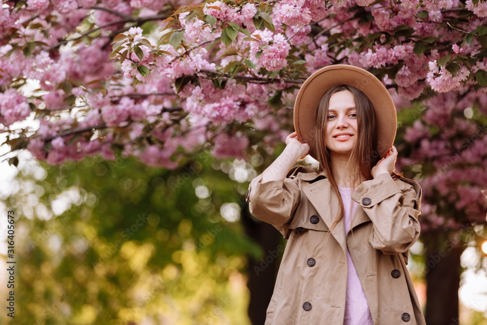 Portrait of young beautiful fashionable girl in hat posing near blooming tree with pink flowers on a sunny day. Spring, girl near a flowering sakura tree. Portrait of young beautiful fashionable girl in hat posing near blooming tree with pink flowers on a sunny day. Spring, girl near a flowering sakura tree.