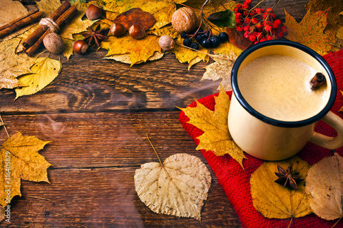 Coffee in an enamel mug with a woolen scarf, leaves, nuts, cinnamon sticks and star anise on wooden background. Autumn cozy background.