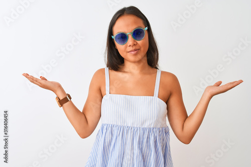 Young chinese woman wearing striped dress and sunglasses over isolated white background clueless and confused expression with arms and hands raised. Doubt concept.