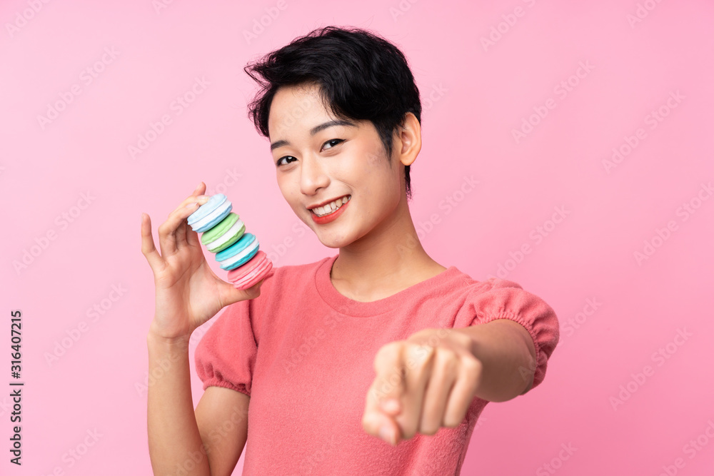 Young Asian girl over isolated pink background holding colorful French macarons and points finger at you Young Asian girl over isolated pink background holding colorful French macarons and points finger at you