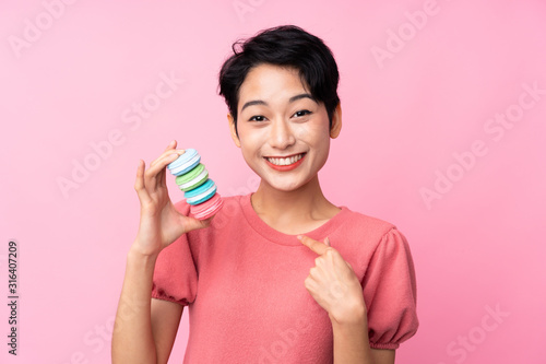 Young Asian girl over isolated pink background holding colorful French macarons with surprise expression