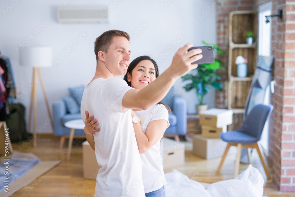 Young beautiful couple standing using smartphone to take a selfie at new home around cardboard boxes Young beautiful couple standing using smartphone to take a selfie at new home around cardboard boxes