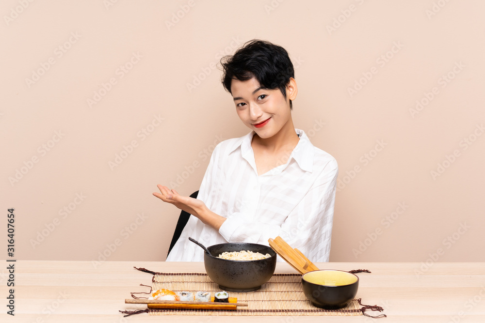 Young Asian girl in a table with bowl of noodles and sushi extending hands to the side for inviting to come Young Asian girl in a table with bowl of noodles and sushi extending hands to the side for inviting to come