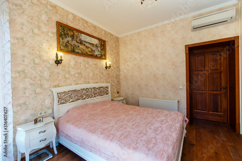 A bedroom in the Provence style. A large white bed with curved legs and back and a pink bedspread. Yellowish Wallpaper with a pattern.