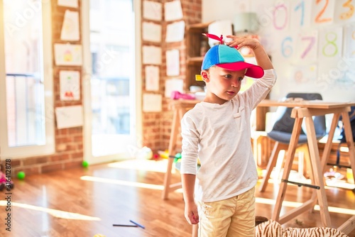 Beautiful blonde toddler wearing fanny colorful propeller cap. Standing around lots of toys at kindergarten