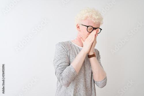 Young albino blond man wearing striped t-shirt and glasses over isolated white background smelling something stinky and disgusting, intolerable smell, holding breath with fingers on nose. Bad smells