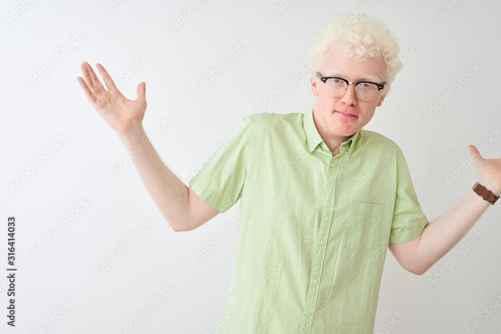 Young albino blond man wearing green shirt and glasses over isolated white background clueless and confused expression with arms and hands raised. Doubt concept. Young albino blond man wearing green shirt and glasses over isolated white background clueless and confused expression with arms and hands raised. Doubt concept.