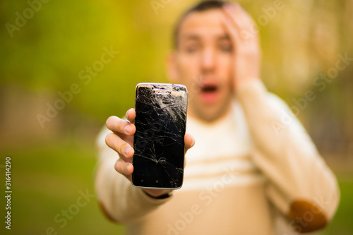 Handsome young man hold broken smartphone screen stressed with hand on head, shocked with shame and surprise face, angry and frustrated.
