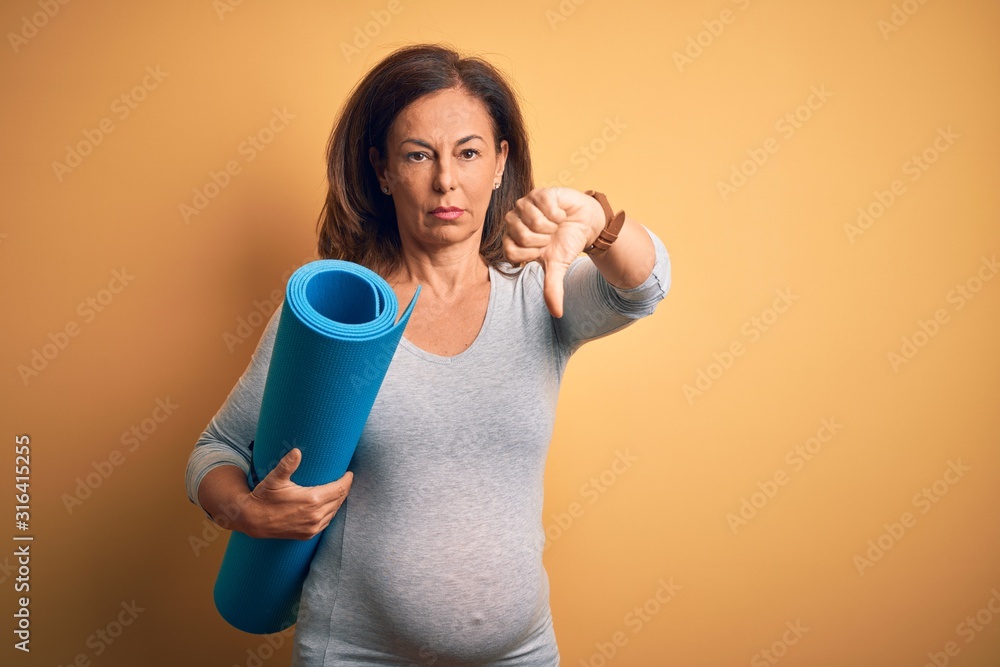 Middle age pregnant woman expecting baby holding yoga mat for healthy lifestyle with angry face, negative sign showing dislike with thumbs down, rejection concept Middle age pregnant woman expecting baby holding yoga mat for healthy lifestyle with angry face, negative sign showing dislike with thumbs down, rejection concept