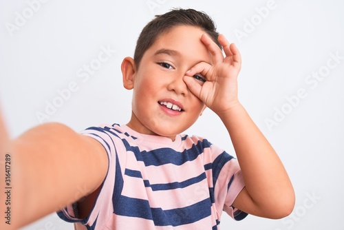 Beautiful kid boy wearing casual striped t-shirt make selfie over isolated white background with happy face smiling doing ok sign with hand on eye looking through fingers