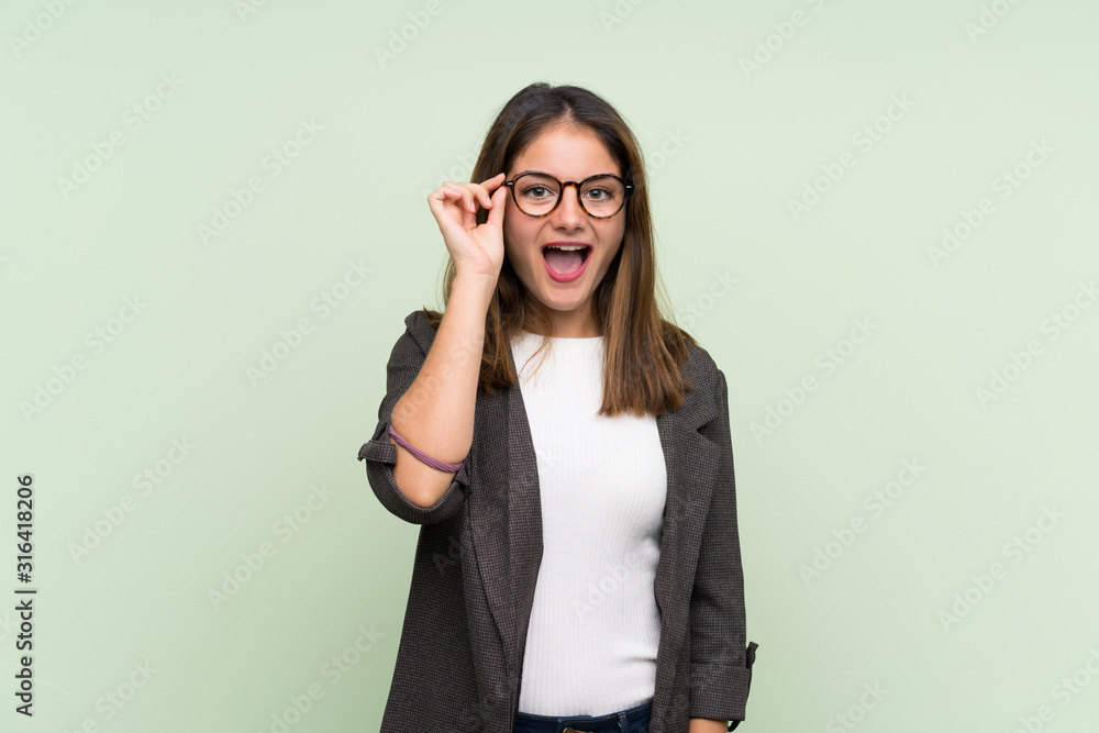 Young brunette girl with blazer over isolated green background with glasses and surprised