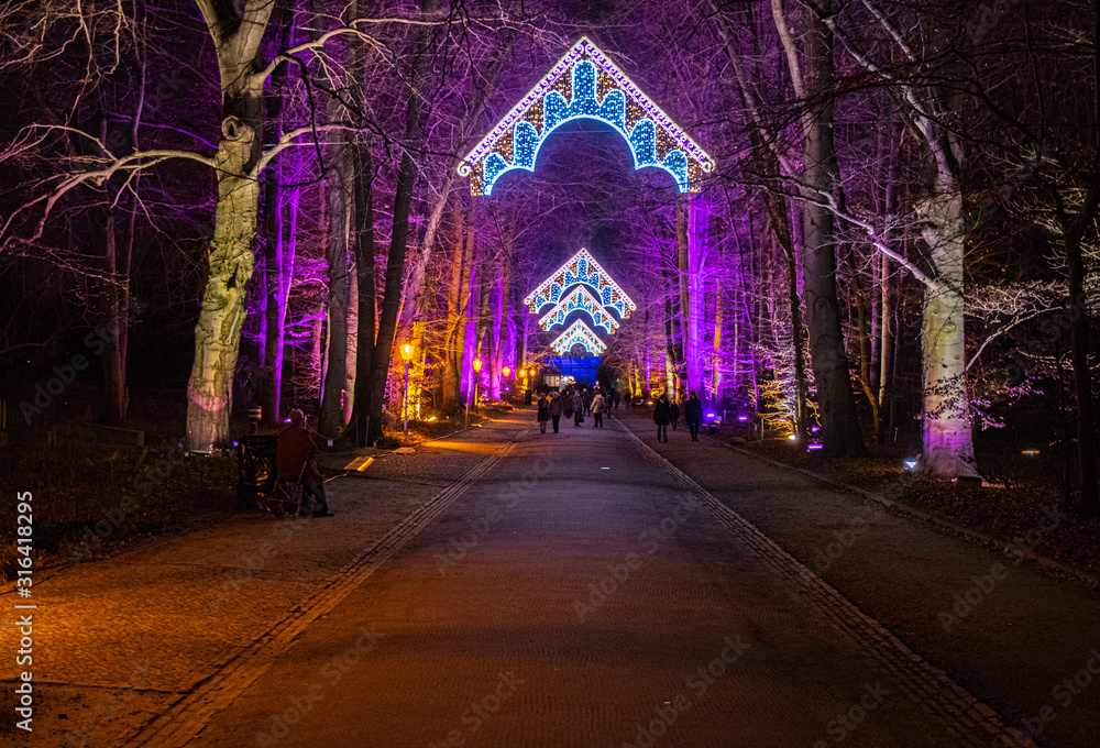 View of entrance to Christmas Garden Berlin. Huge colorful illuminated trees by sides of wide road and ornate lighted arches above. Diminishing perspective winter landscape in night