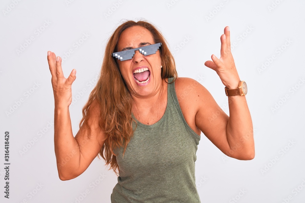 Middle age mature woman wearing thug life sunglasses over isolated background crazy and mad shouting and yelling with aggressive expression and arms raised. Frustration concept. Middle age mature woman wearing thug life sunglasses over isolated background crazy and mad shouting and yelling with aggressive expression and arms raised. Frustration concept.