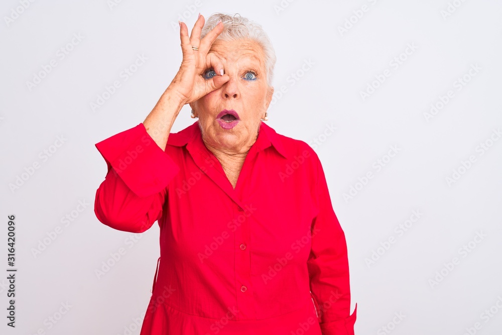 Senior grey-haired woman wearing red casual shirt standing over isolated white background doing ok gesture shocked with surprised face, eye looking through fingers. Unbelieving expression. Senior grey-haired woman wearing red casual shirt standing over isolated white background doing ok gesture shocked with surprised face, eye looking through fingers. Unbelieving expression.