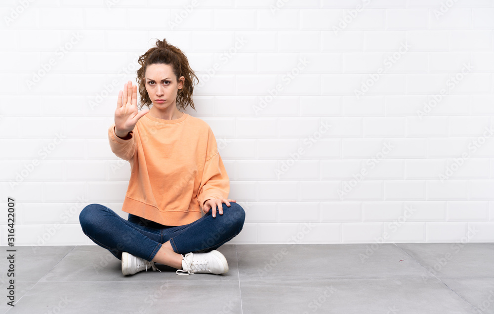 Fototapeta premium Young woman with curly hair sitting on the floor making stop gesture with her hand